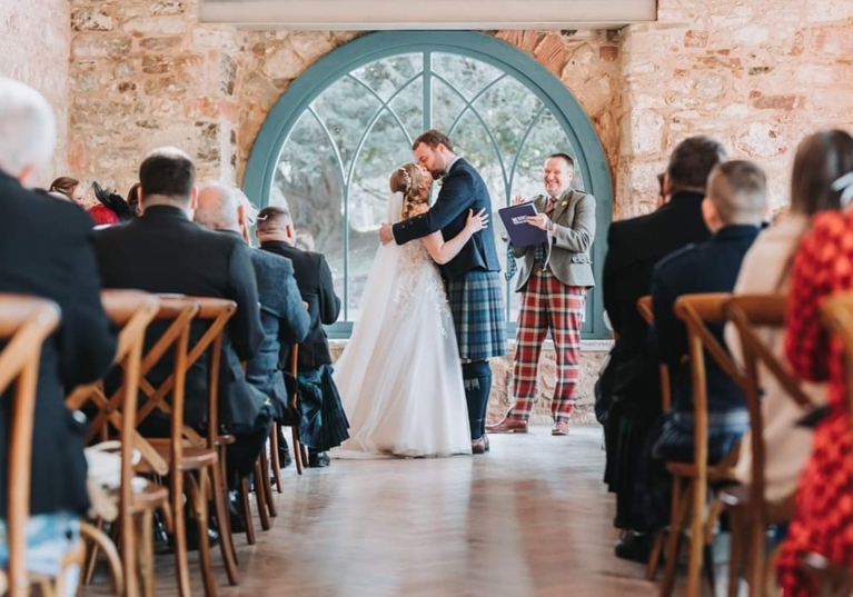 Bride and groom kiss at altar with celebrant smiling and clapping in background